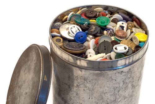 A cylindrical metal container, possibly a tin or waste bin, filled with an assortment of various plastic buttons in multiple shapes, sizes, and colours, including black, white, blue, red, yellow, and green. The container has a slightly worn, matte finish with visible scratches and a partially opened metal lid placed on the ground nearby. The objects inside are tightly packed, and the background is plain white, indicating the image focuses solely on the container and its contents. This scene reflects rubbish or waste collection, relevant to garden clearance and general rubbish removal services in the Chigwell area, as provided by Garden Clearance Chigwell.