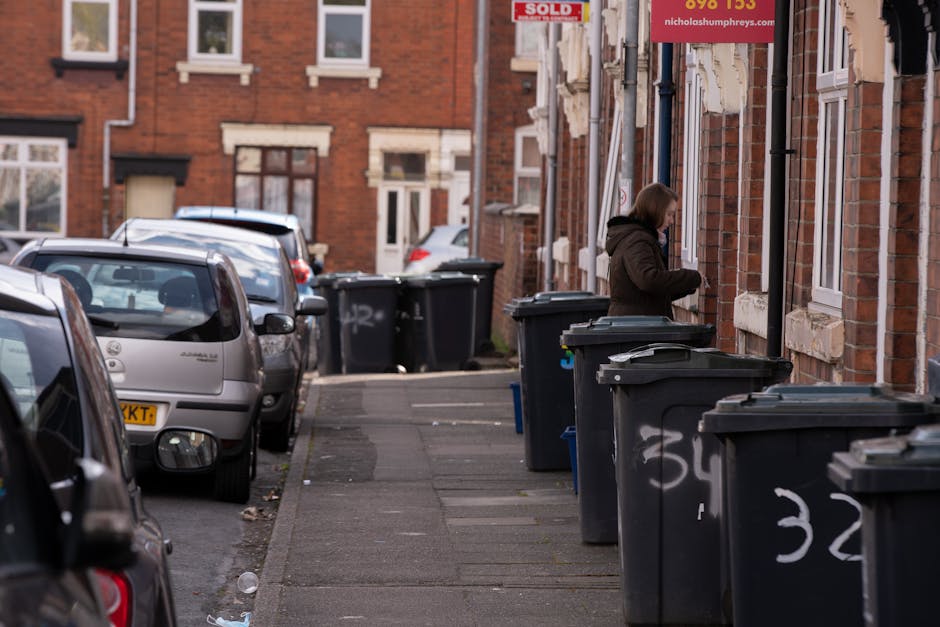A street scene featuring a white waste collection truck with a rusted and worn metal rear compartment actively engaged in rubbish collection on a narrow cobblestone road. The truck is positioned paral