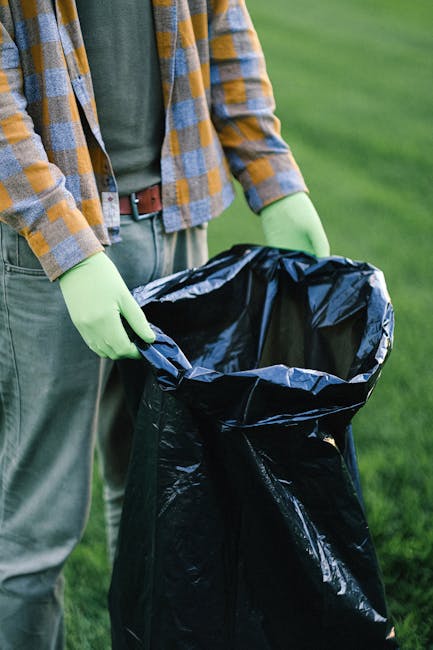 Close-up of a person wearing a plaid shirt and grey trousers, holding open a large black rubbish bag lined with a silver interior, with their hands protected by bright green gloves. The background sho